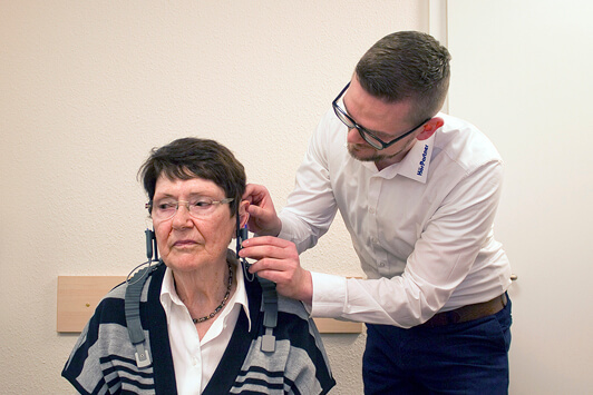 Ein Hörakustiker passt einer Seniorin in einer Praxis ein Hörgerät an.